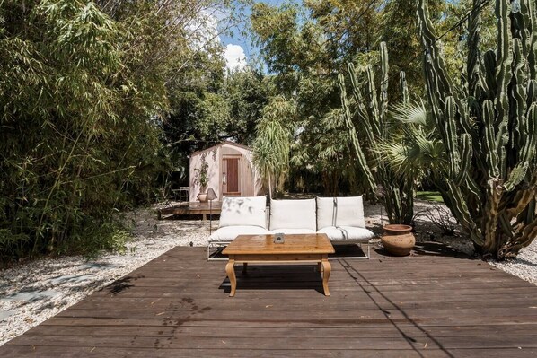 Una terraza de madera con un sofá blanco y una mesa de madera, rodeada de cactus y plantas de bambú.