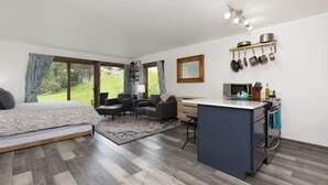 Un dormitorio moderno con una cama grande, un área de estar con sofás, una pequeña cocina con barra y vistas al exterior.