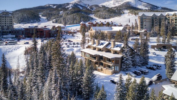 Un complejo turístico cubierto de nieve con varios edificios, incluyendo una cabaña principal con tejado nevado y un estacionamiento con varios automóviles.
