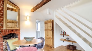 A dining area with a brick fireplace, wooden ceiling beams, and a staircase leading to a loft. - Wherry Cottage (Manningtree)