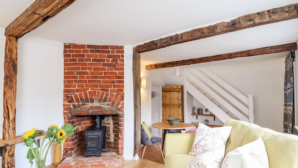 A cozy living room with a brick fireplace, wooden beams, a sofa with cushions, a vase with sunflowers, and a staircase in the background. - Wherry Cottage (Manningtree)