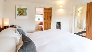 A bedroom with a large bed, a bedside table with a lamp, a chair, a wooden door, and a framed picture on the wall. - Wherry Cottage (Manningtree)