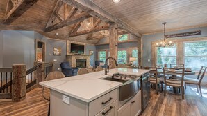 Un intérieur rustique avec des poutres apparentes, une cheminée en pierre, un coin repas doté d’un grand îlot central et une vue sur la verdure par les fenêtres.