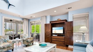 Ein Wohnzimmer mit Glastisch, zwei gemusterten Sofas, einem TV-Schrank mit Flachbildfernseher und Blick auf einen Balkon mit Gartenmöbeln.