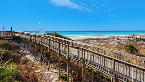 Ein hölzerner Steg führt zu einem Strand unter blauem Himmel.