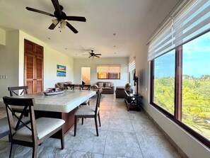 A spacious living area with a dining table, seating, a ceiling fan, and a large window offering a view of greenery.