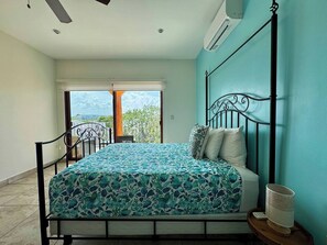 A bedroom with a bed, a nightstand, a wall-mounted air conditioner, and a view of the outside.