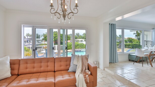 Ein großzügiges Wohnzimmer mit Ledersofa, einer Chandelüster und großen Fenstern mit Blick auf einen Pool und Grünflächen.