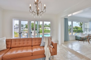A spacious living room with a leather sofa, a chandelier, and large windows offering a view of a swimming pool and greenery. - Waterfront w/ Dock | Pool, Kayaks & Dolphin Sightings (Bradenton)