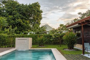 A swimming pool surrounded by lush greenery, a wooden deck, and a modern building with a sloped roof.
