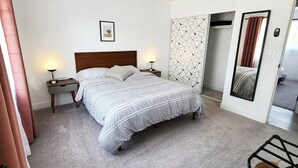 Une chambre à coucher comprenant un lit, des tables de chevet, un miroir fixé au mur et une porte donnant sur une autre pièce.