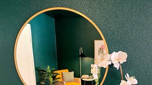 Une pièce avec un miroir rond, une étagère en bois, une chaise jaune, une petite table et un pot d’orchidée.