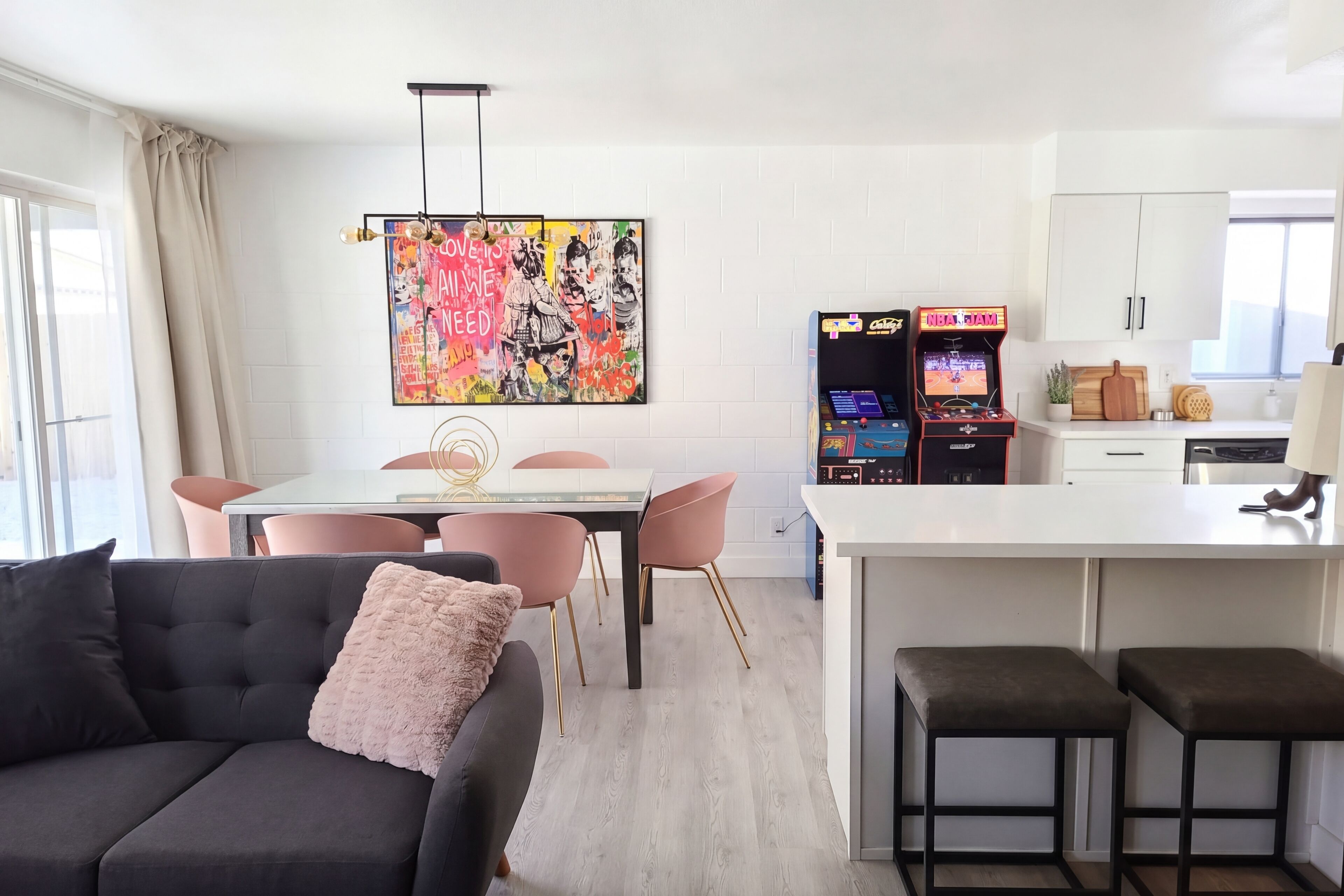 A modern kitchen with white cabinets, a dining area with a glass table and pink chairs, and two arcade games.