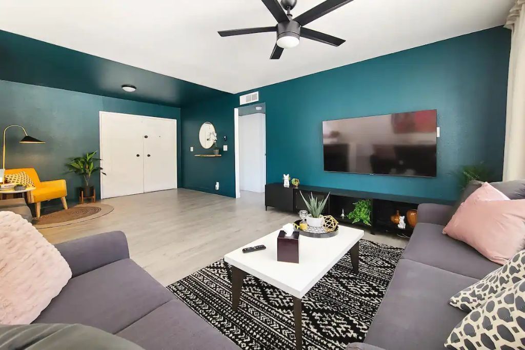 Un salon moderne avec des murs de couleur turquoise, un téléviseur à écran plat, un ventilateur de plafond et un canapé agrémenté d’un tapis à motifs.