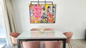 Un coin repas comprenant une table à plateau en verre, quatre chaises roses aux pieds dorés et un tableau abstrait aux couleurs vives accroché au mur.