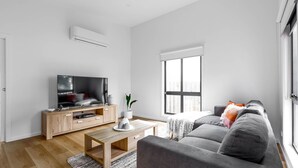 Een moderne woonkamer met een flatscreen televisie op een houten tv-meubel, een grijze bank, een salontafel en een groot raam dat natuurlijk licht binnenlaat.