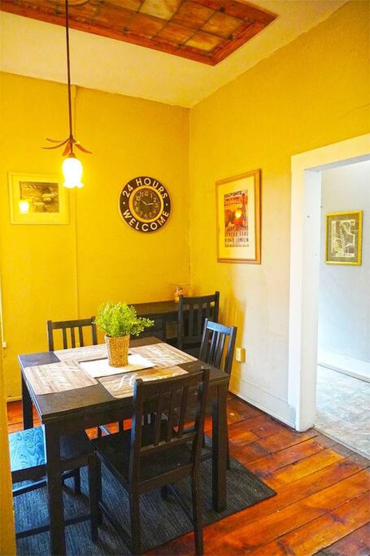 A dining area with a wooden table and chairs, a hanging light fixture, a clock, and framed pictures on the walls.
