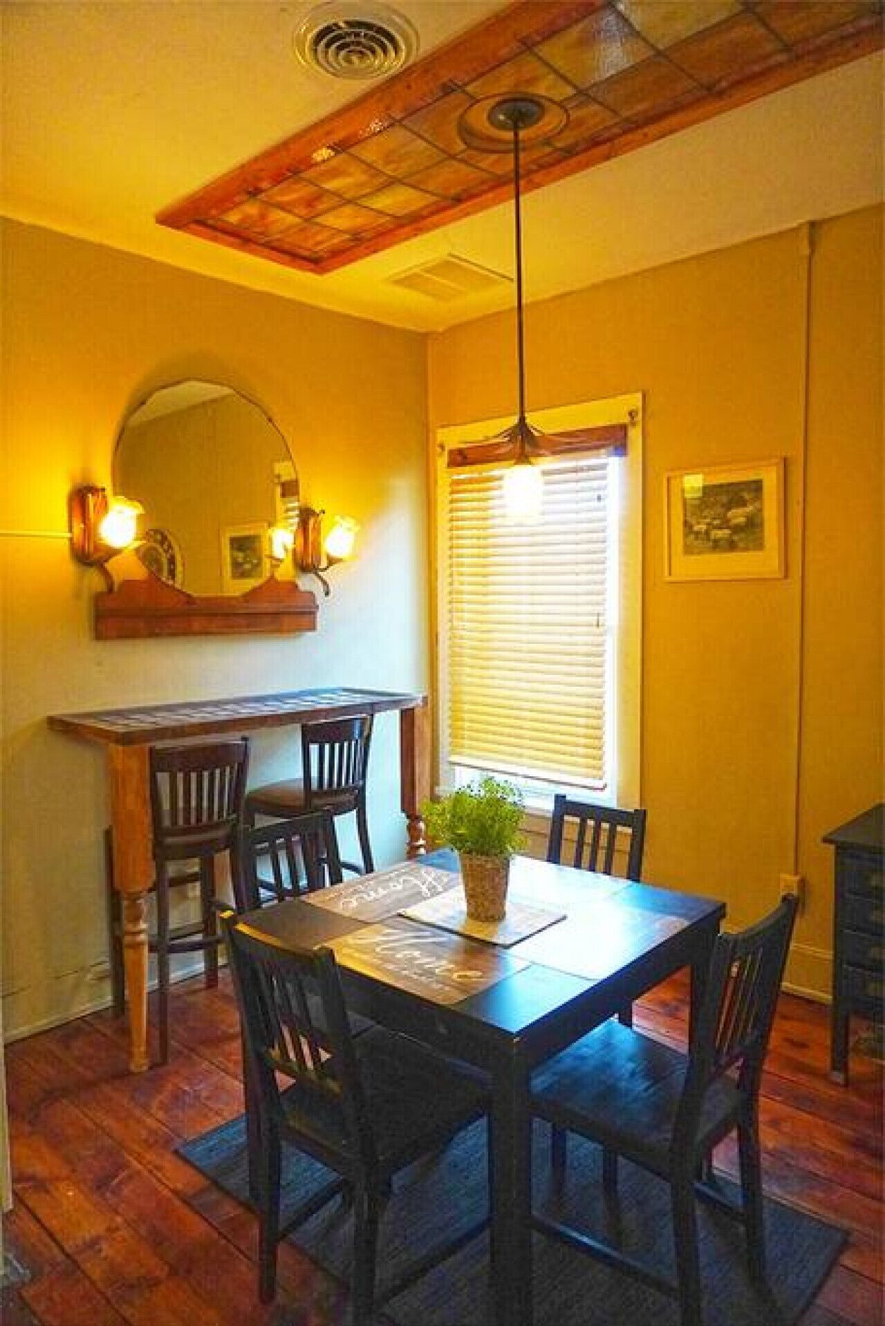 A dining area with a wooden table and chairs, a hanging light fixture, a window with blinds, and a mirror on the wall.