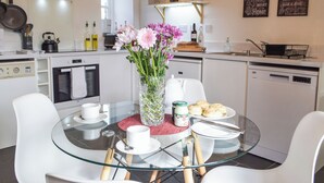 A modern kitchen with a glass dining table, white chairs, a vase of flowers, and various kitchen appliances.