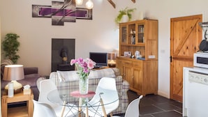 A cozy living room with a glass table, white chairs, a plaid sofa, a wooden cabinet, and a kitchen area with a stove.
