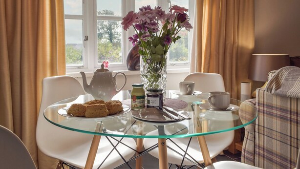 A glass table with a teapot, a vase with flowers, biscuits, and a jar of cream.