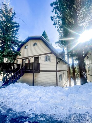 A two-story house with a wooden staircase leading to a deck, surrounded by snow and tall trees.