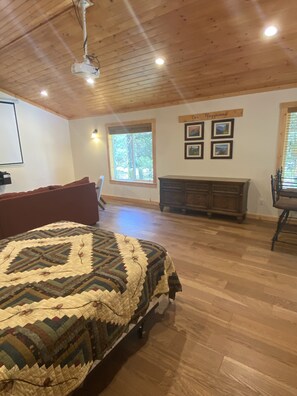 A spacious room with a large bed, a wooden ceiling, a window with a view of trees, and a wall-mounted TV.