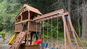 A wooden play structure with a slide and swings, set on a gravel surface with trees in the background. - Kid Friendly Cabin with Mother-in-law Suite and Hot Tub! (Ronald)