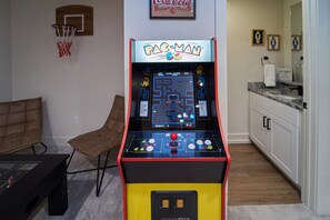 Máquina de arcade Pac-Man numa sala com um cesto de basquete, um sofá e uma área de cozinha.