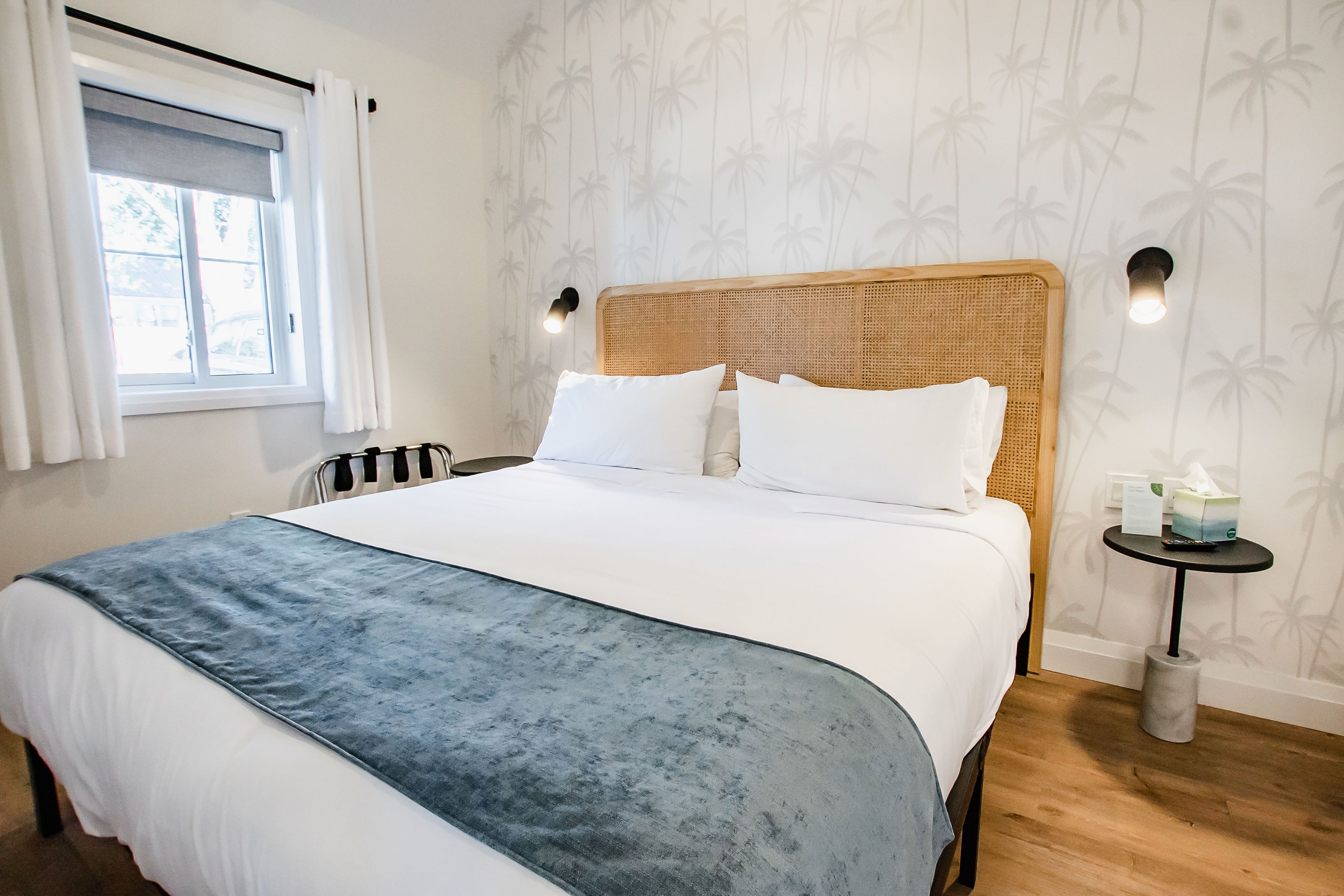 A neatly made bed with white linens and a blue accent, a wicker headboard, and a small bedside table with a tissue box.
