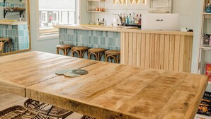 L’intérieur d’un restaurant présente une grande table en bois, des chaises en osier, des lampes en osier suspendues au plafond et un comptoir orné d’une enseigne au néon.