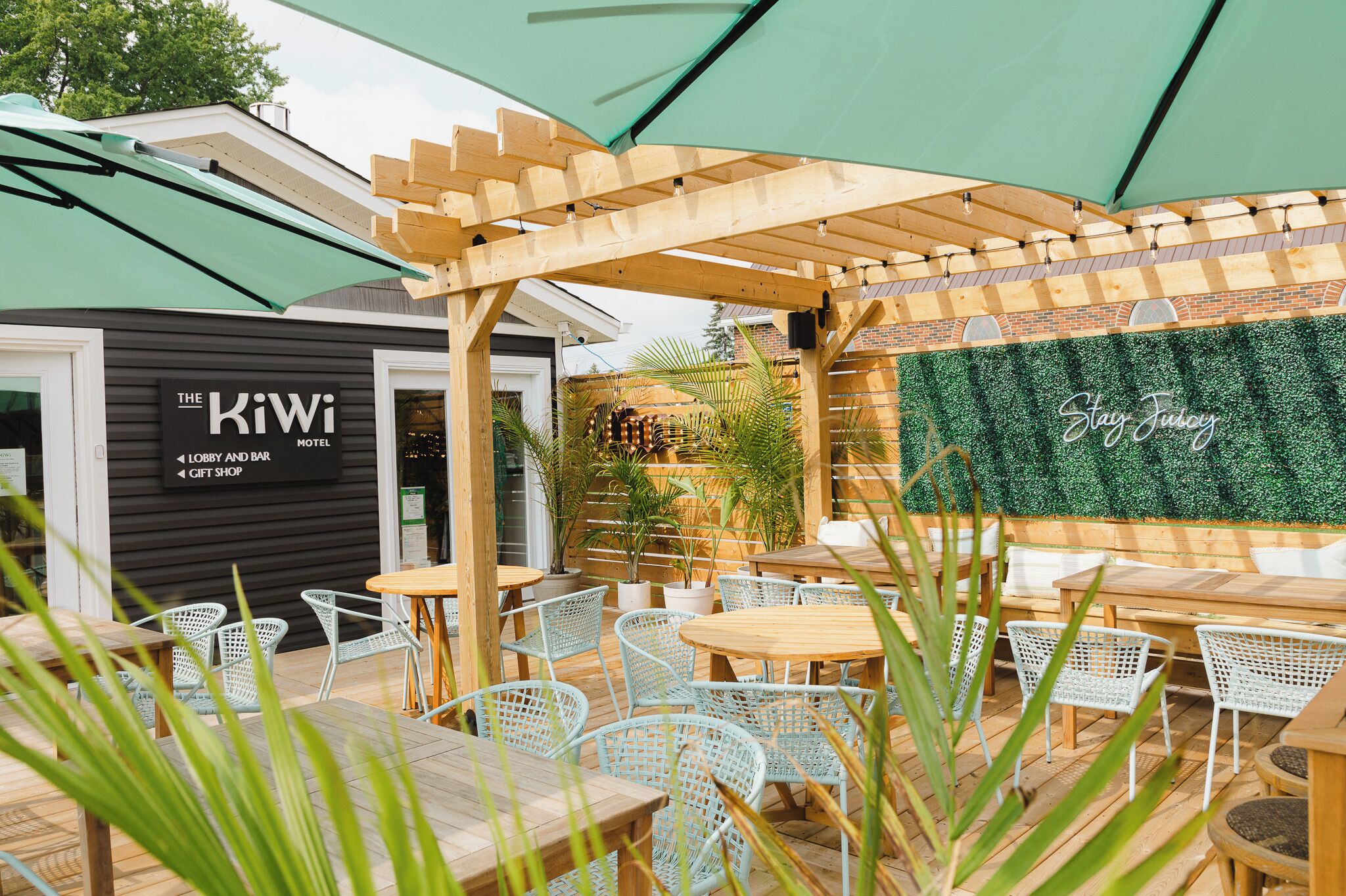 Un espace extérieur aménagé avec des tables en bois et des chaises en osier blanc, une pergola en bois et une enseigne indiquant « The Kiwi Motel », avec les mentions « réception et bar » et « boutique de souvenirs ».