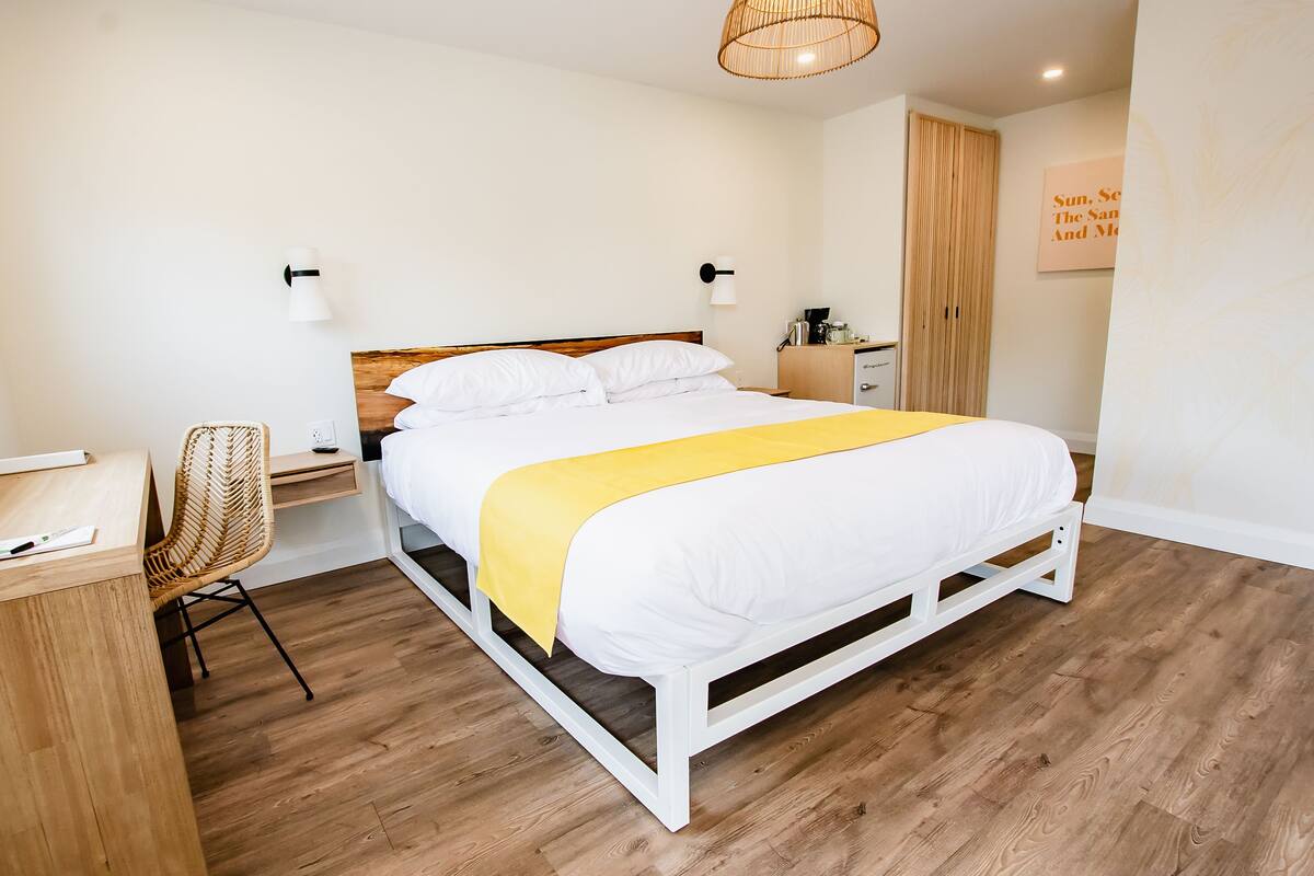 Een keurig opgemaakt slaapkamer met een bed, nachtkastje, stoel en een wandlamp.