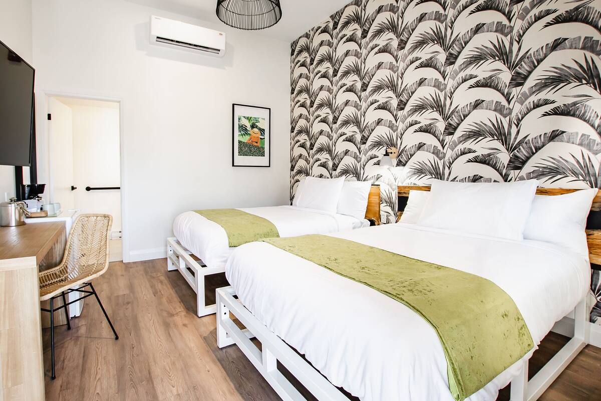 Een hotelkamer met twee bedden, een bureau en een stoel. Er is een wandairco en een ingelijst kunstwerk aan de muur.