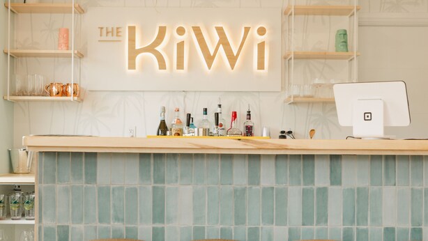 Un espace bar agrémenté d’une enseigne indiquant « The Kiwi », avec des suspensions en osier, un mur recouvert de carreaux et des tabourets en bois.