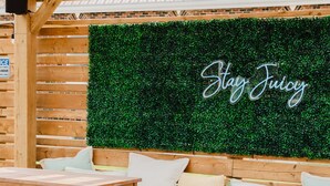 Un espace extérieur aménagé avec des tables et des chaises en bois, un mur végétal orné de l’inscription « Stay Juicy », et une structure de pergola en bois.