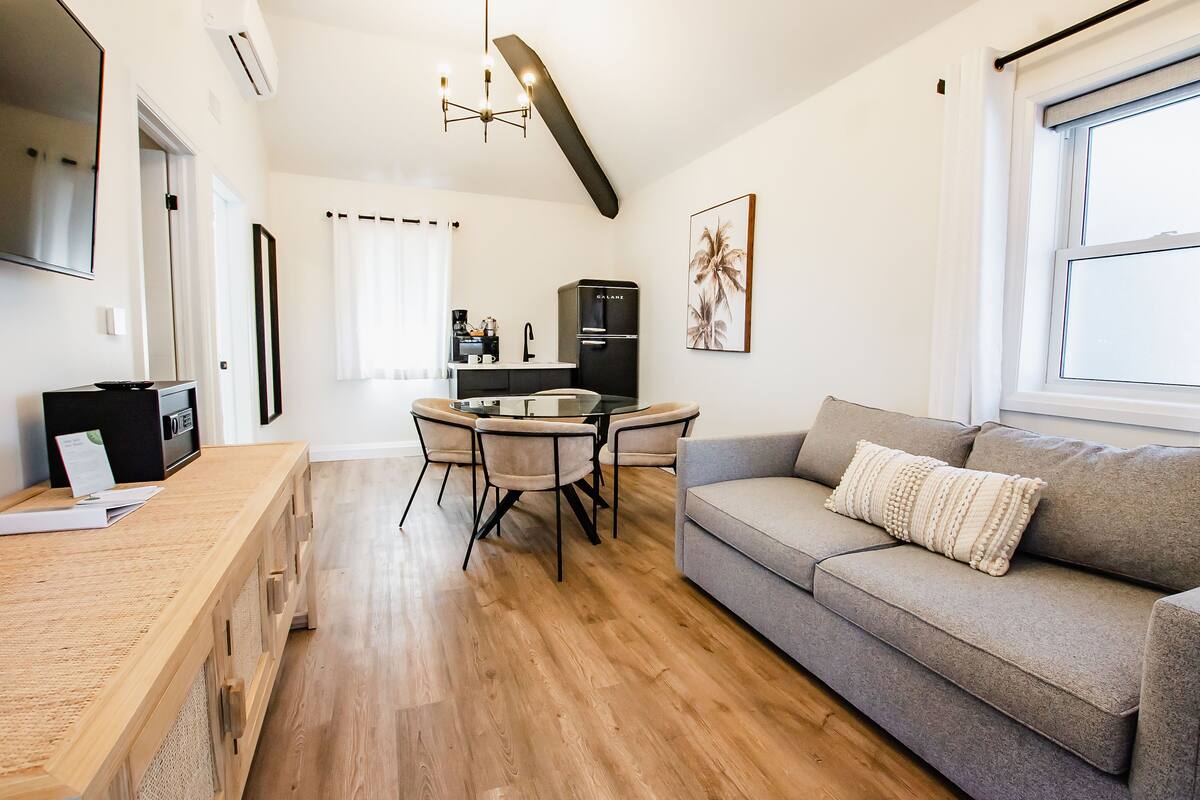 Een moderne woonkamer met een grijze bank, een houten salontafel en een eethoek met een glazen tafel en stoelen.
