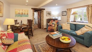 A cozy living room with a blue sofa, a wooden coffee table with a fruit bowl, a patterned rug, and a dining area with a wooden table and chairs. - Southcott Farm Cottage (Honiton)