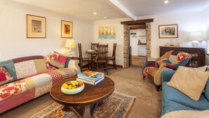 A cozy living room with a sofa, a coffee table with a fruit bowl, a dining table with chairs, and a kitchen area visible through an open doorway. - Southcott Farm Cottage (Honiton)