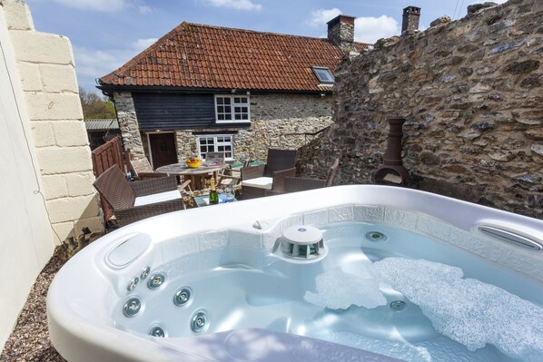 A hot tub in a backyard with a stone wall, a wooden deck, and a house with a tiled roof. - Southcott Farm Cottage (Honiton)