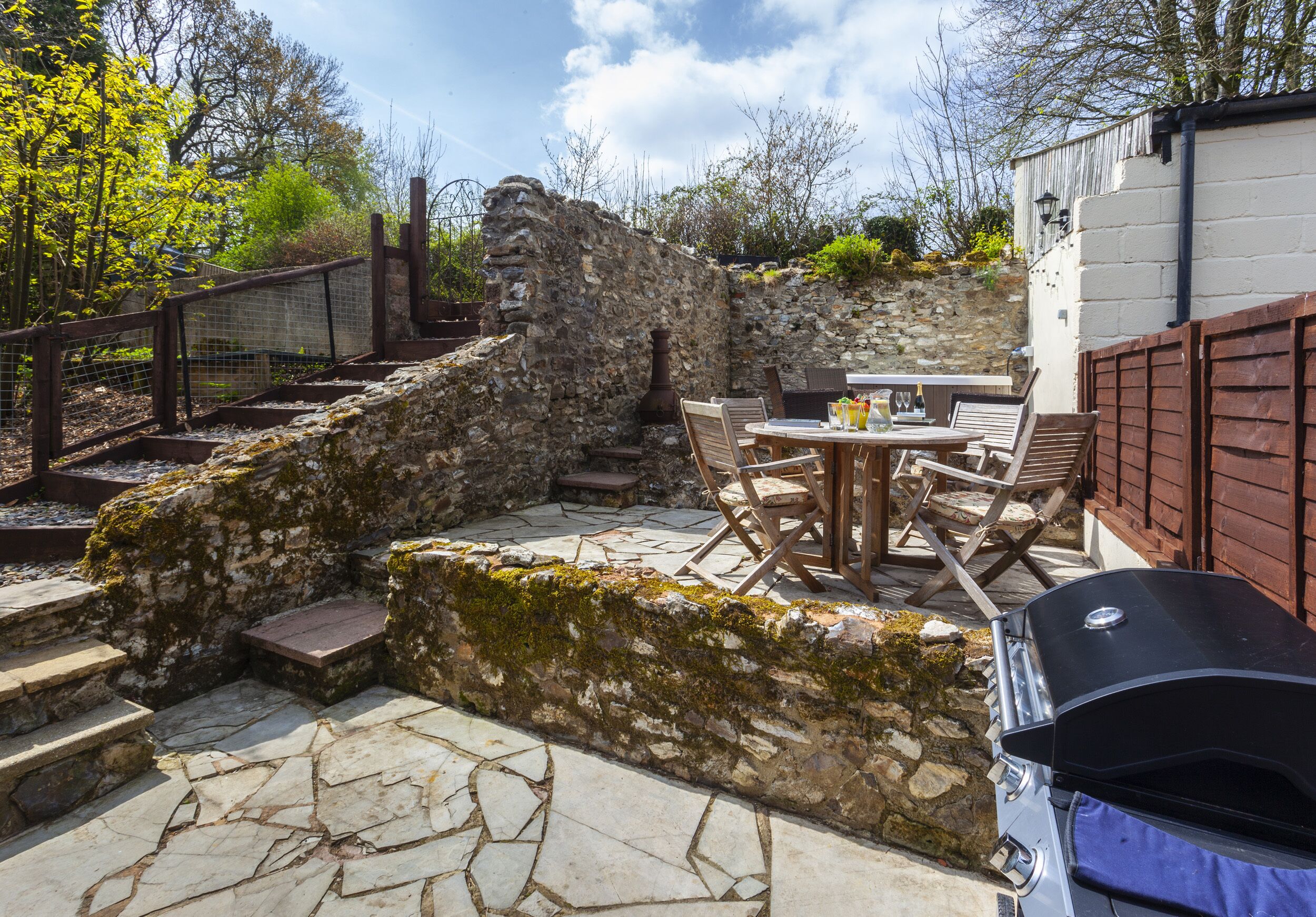 Un patio de piedra con una mesa y sillas, una parrilla de barbacoa y un muro de piedra con escalones que llevan a una terraza de madera.