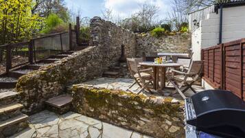 Un patio de piedra con una mesa y sillas, una parrilla de barbacoa y un muro de piedra con escalones que llevan a una terraza de madera.