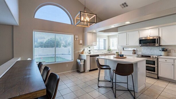 A modern kitchen with white cabinetry, a central island with bar stools, a large window with blinds, and a skylight.