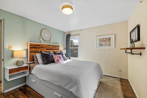A bedroom with a bed, bedside table, lamp, wall art, and a window.