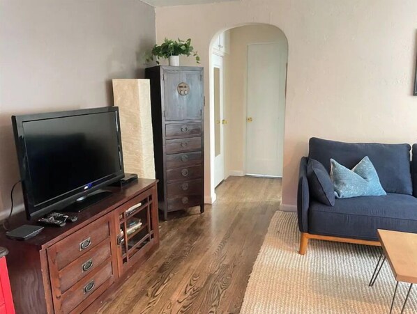 A living room with a flat-screen TV on a wooden cabinet, a dark blue sofa with cushions, and a wooden floor.