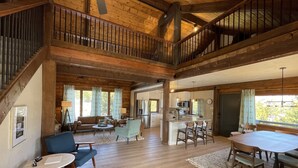 A spacious living area with a high ceiling, wooden beams, a staircase, a kitchen with a central island, and a dining area with a table and chairs. - Midcentury Mountain Cabin, complete with hot tub, game room and amazing VIEWS!! (Grand Lake)