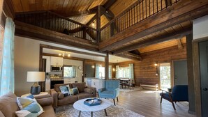 A spacious living area with a high ceiling, wooden beams, a sofa set, a coffee table, and a view of the kitchen and dining area. - Midcentury Mountain Cabin, complete with hot tub, game room and amazing VIEWS!! (Grand Lake)