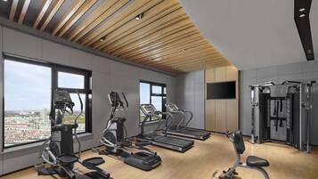 A modern gym with various cardio machines, a TV mounted on the wall, and large windows offering a city view.