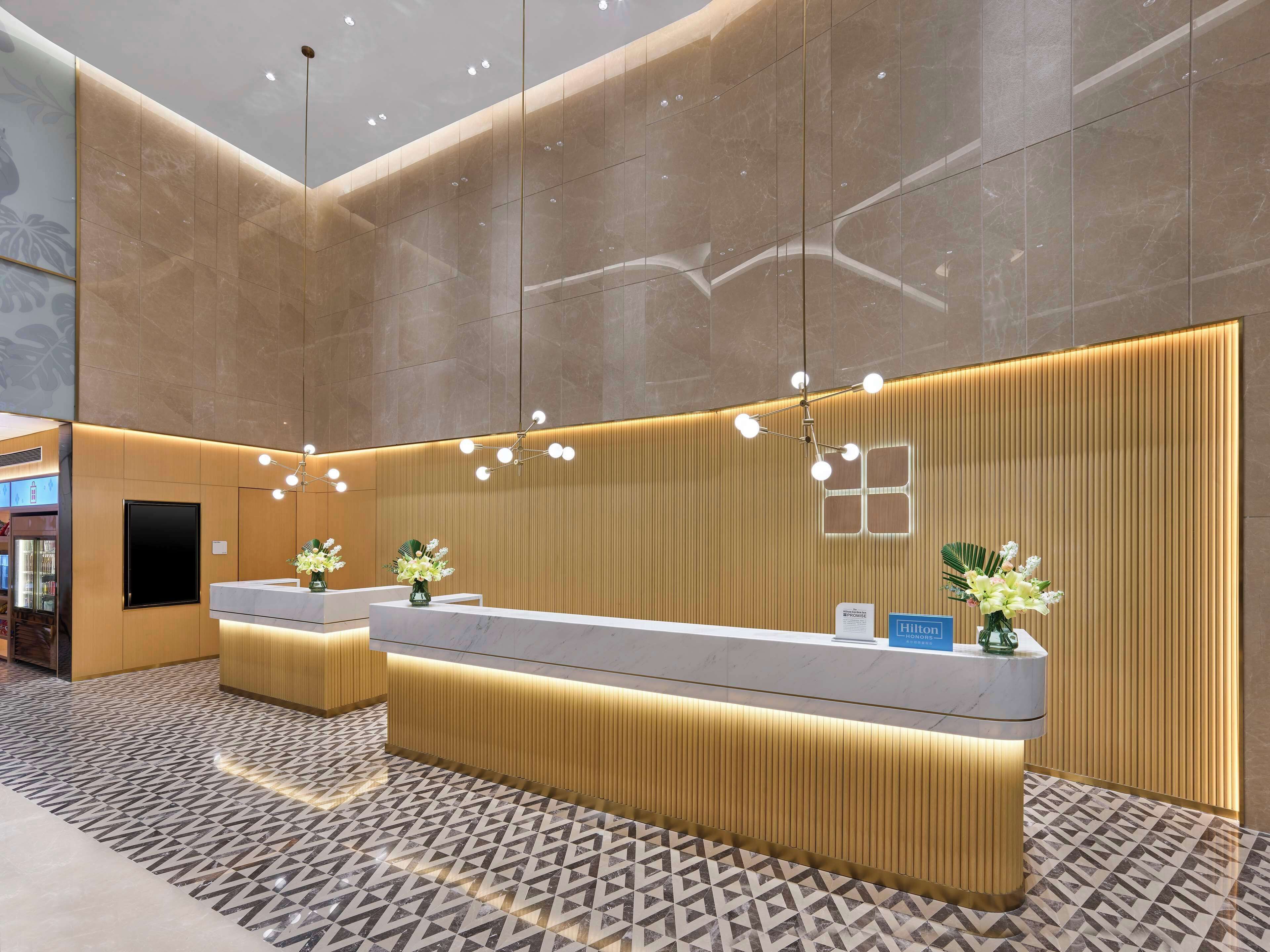 Un área de recepción de hotel moderna con un mostrador central, dos mostradores laterales y un espejo grande.