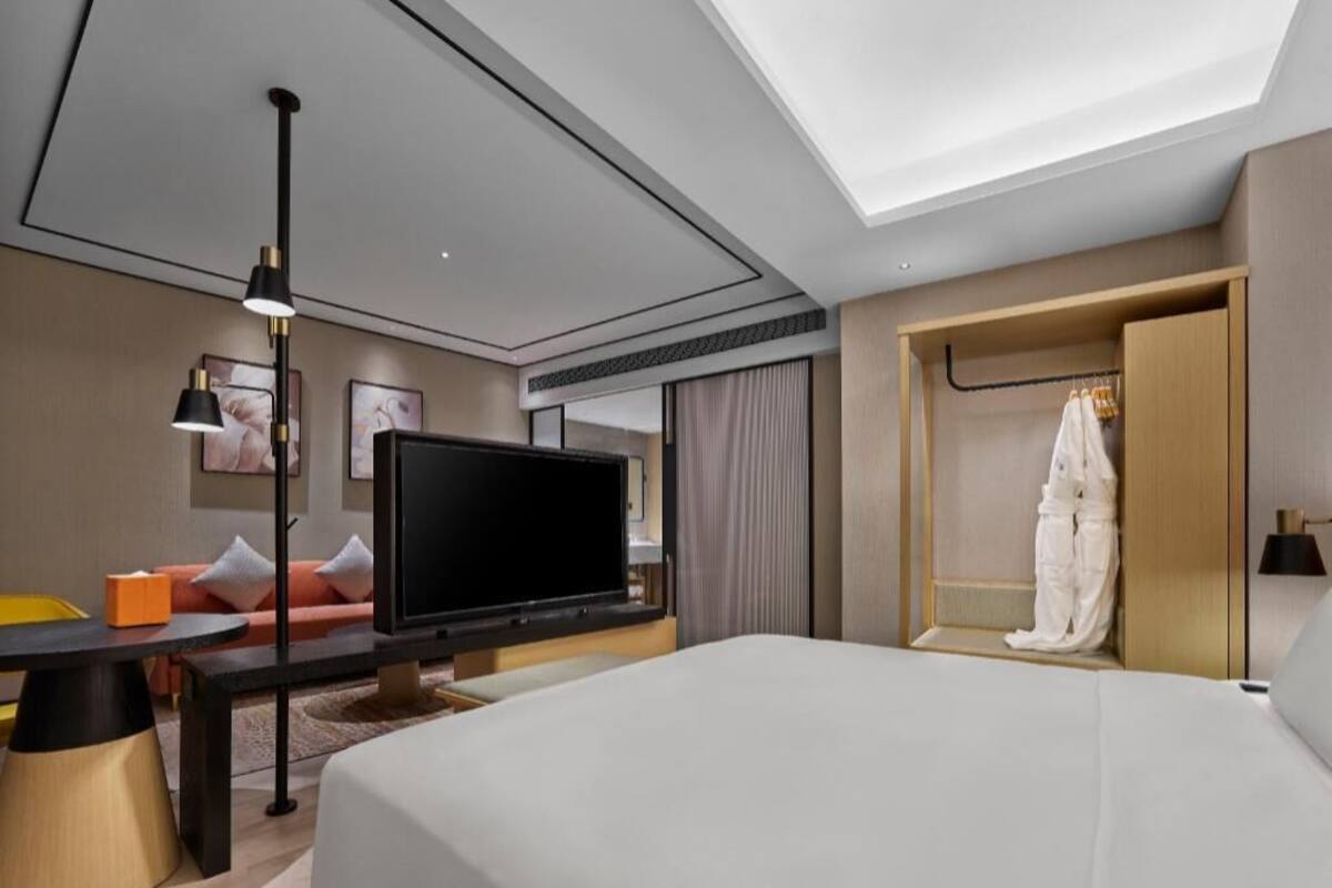A modern hotel room with a bed, a flat-screen TV, a desk with a lamp, and a wardrobe.